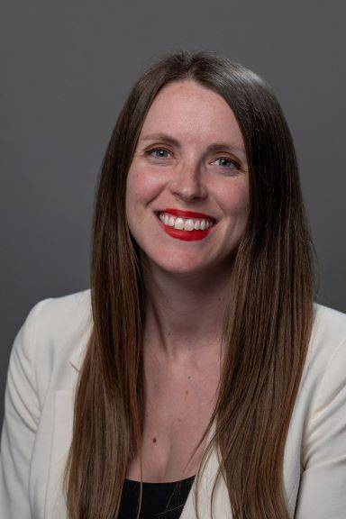 A formal headshot of Michele, who has long brown hair and is wearing a white blazer